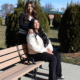 Two women sit on a park bench surrounded by neatly trimmed bushes, enjoying a sunny day outdoors. One has her hand on the other's shoulder.